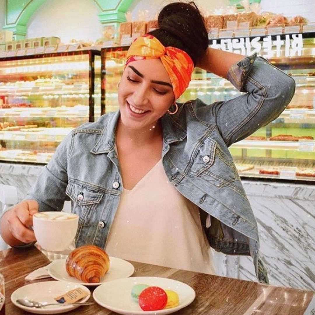 Woman wearing a Blush Cosmo scented scarf styled as a headband in a café setting.